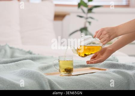Frau gießt heißen Kräutertee aus Glas-Teekanne in Becher auf Holztablett in gemütlicher Schlafzimmereinstellung, in einem gemütlichen Schlafzimmer mit einer weichen Decke auf dem Bett, Stockfoto