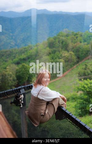 Glückliche lächelnde asiatische Frau sitzt auf der Holzterrasse mit Blick auf die grünen Wälder Bergkette während des Sommerreiseurlaubs und genießt friedliche Natur Stockfoto