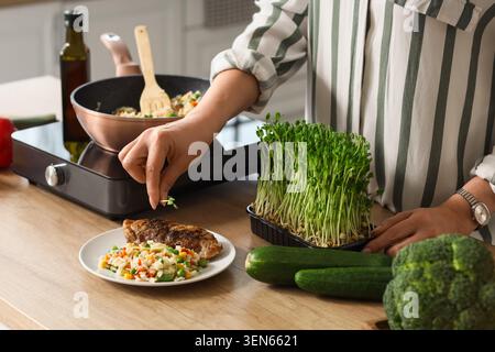 Frau garniert gesundes gegrilltes Hühnchen und Gemüsereis mit frischem Mikrogrün für nahrhafte Hausmannskost, die Mahlzeit wird auf einem modernen Industriezweig zubereitet Stockfoto