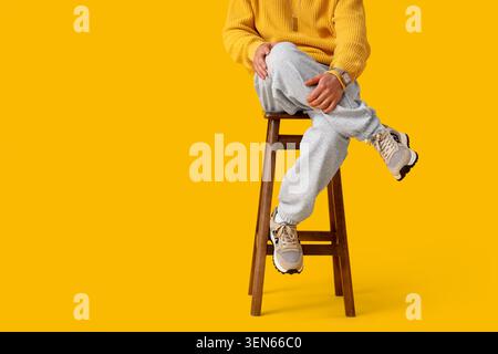 Legere junge Person in gelbem Pullover und grauer Jogginghose, die auf Holzhocker sitzt, moderne Streetwear-Mode Stockfoto