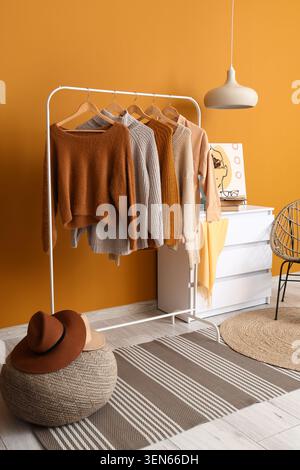 Herbstliche Pullover und Cardigans auf Kleiderständer in gemütlichem Zimmer mit orangefarbenen Wänden und moderner Einrichtung, die warme orangefarbene Wand ergänzt die moderne Einrichtung Stockfoto