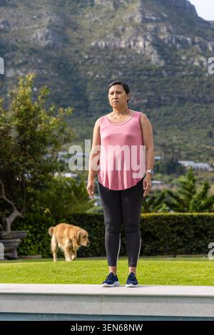 Reife Frau, die auf Steinrand im Garten steht, trägt rosa Top Leggings Armbanduhr Hund läuft Stockfoto
