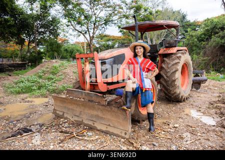 Traktor Landwirt Frau Bauernhof Klinge Schlammstiefel Hut ländliches Feld, Frau Landwirt in buntem Hemd stehend bei rotem Traktor mit Frontmesser auf schlammiger ländlichem Bauernhof Stockfoto