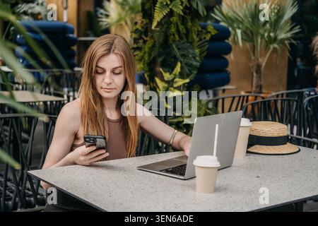 Die junge Frau sitzt mit Smartphone und Laptop am Kaffeetisch im Freien. Plattformübergreifende Kommunikation, synchronisierte digitale Aufgaben, Multitasking für Frauen Stockfoto