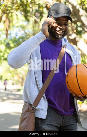 Erwachsener afrikanischer Mann mit schwarzer Kappe, grauem Hoodie, Basketball in der Hand, Smartphone auf dem Weg Stockfoto