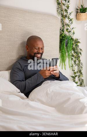 Reifer afroamerikanischer Mann, der auf dem Bett liegt und ein Smartphone hält, mit einem dunkelgrauen Hemd Stockfoto