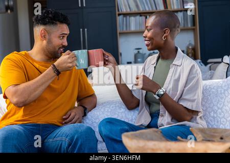 Diverse Paare sitzen auf dem Sofa zu Hause und knacken Keramikbecher, Mann orange T-Shirt, Frau beige Overshirt Stockfoto