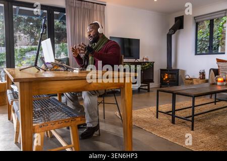 Afroamerikaner sitzt am Esstisch mit Schal und Headset, spricht mit Laptop ins Mikrofon Stockfoto