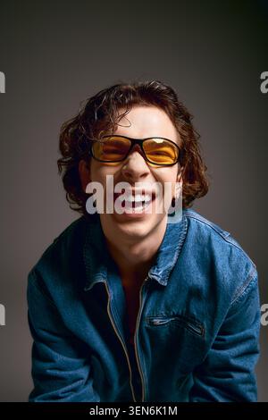 Ein junger Mann mit lockigen Haaren lächelt glücklich in trendiger Sonnenbrille und Outfit. Stockfoto
