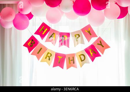 Leuchtend rosa und weiße Ballons umrahmen elegant ein schönes Happy Birthday-Banner mit glänzenden goldenen Schriftzug, das vor einem hellen, sonnendurchfluteten Wind steht Stockfoto