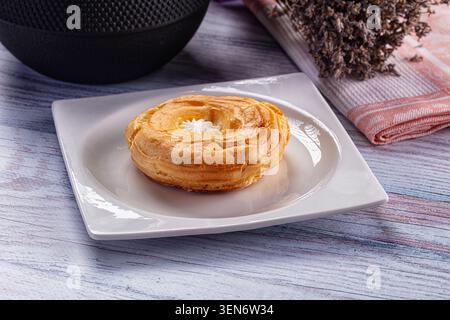 Goldener brauner Choux-Gebäckring mit Creme auf weißem quadratischem Teller. Schwarze Schüssel mit getrocknetem Lavendel im Hintergrund auf rustikalem blauem Holztisch Stockfoto