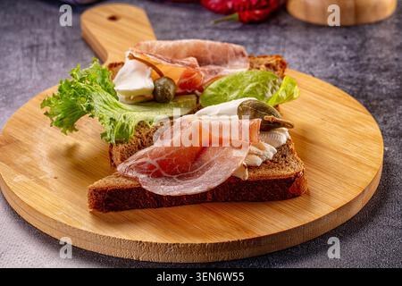 Offenes Sandwich mit Schinken auf Holzbrett. Zwei Scheiben dunkles Brot mit geräuchertem Schinken, Weichkäse, Salat und Kapern Stockfoto