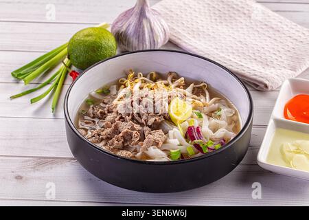 Traditionelle vietnamesische Rindfleischsuppe serviert in einer schwarzen Schüssel mit Reisnudeln zarte Rindfleischscheiben Sojasprossen Limettenkeil grüne Zwiebeln Knoblauch und Di Stockfoto