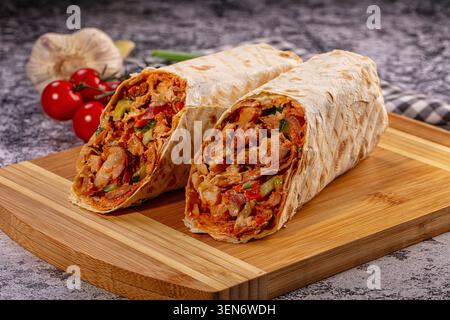 Zwei Hälften einer herzhaften Hühnchen-Shawarma-Wrap gefüllt mit gegrilltem Fleisch, Paprika und Sauce, serviert auf einem Holzbrett mit frischen Tomaten und Knoblauch Stockfoto