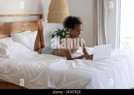 Afroamerikanerin, die auf dem Bett liegt und weiße Kissen auf silbernem Laptop in einem modernen Schlafzimmer spielt Stockfoto