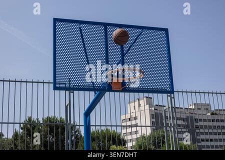 Basketball fliegt auf einem urbanen Spielplatz mit Stadtgebäuden dahinter auf einen blauen Basketball Stockfoto