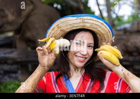 Frau Bananenhut Lächeln Schlamm Obst Porträt tropische traditionelle Bauernhof junge asiatische Frau mit Strohhut und roter traditioneller Kleidung lächelt während des Haltens Stockfoto