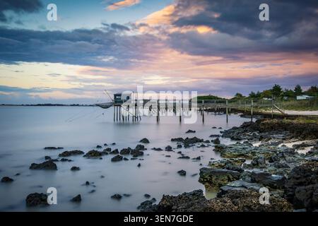 Eine ruhige Küstenszene mit hölzernem Pier, der sich bei Sonnenuntergang über ruhigem Wasser erstreckt, mit dramatischen pastellfarbenen Wolken und zerklüfteter felsiger Küste, die einen friedlichen Frieden schaffen Stockfoto