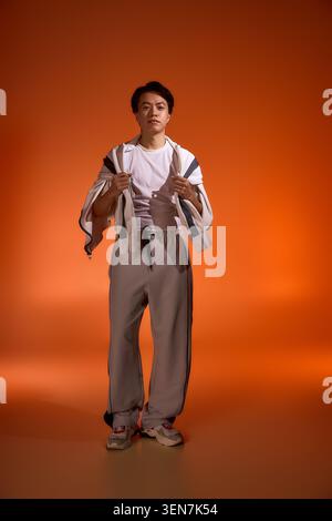 Ein junger asiatischer Mann zeigt sein trendiges Outfit in einem lebendigen Studio mit warmer Beleuchtung. Stockfoto
