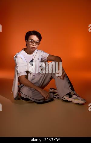 Stilvoller junger asiatischer Mann sitzt souverän in einem Studio mit einem warmen orangen Hintergrund. Stockfoto