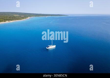 Eine Drohne aus der Luft, die ein einsames weißes Segelboot auf dem pulsierenden blauen Wasser der Adria in der Nähe der Küste der Insel Silba, Kroatien, fängt Stockfoto