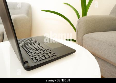 Fernarbeit von zu Hause aus. Laptop auf einem weißen Couchtisch in der Nähe des Sessels. Der Arbeitsplatz von Freiberuflern. Stockfoto