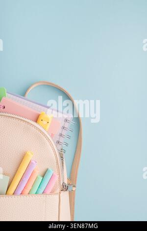 Der Rucksack aus beigefarbenem Kunstleder lässt sich öffnen, um spiralförmige Notizbücher, Textmarker in Pastellfarben, Stifte und ein niedliches Lesezeichen für Schweine auf weichem blauem Hintergrund zu entdecken Stockfoto
