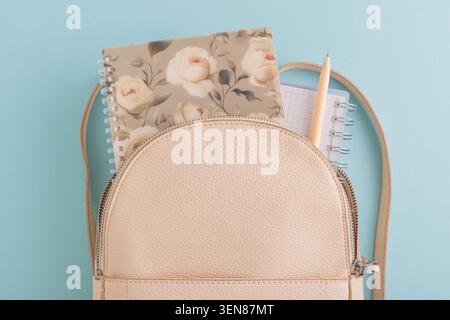 Beige, modische Rucksacköffnung, die ein Notizbuch mit Blumenmuster, ein kleineres Notizbuch mit Gitter und einen beigefarbenen Stift zeigt, der Schule, Universität und Studium repräsentiert Stockfoto