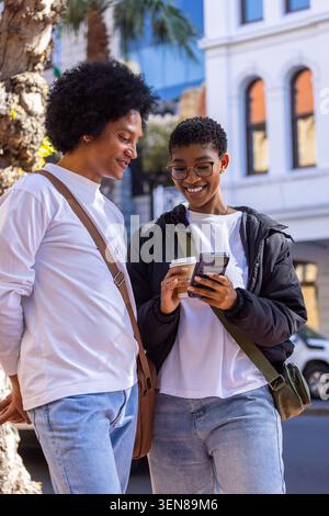 Paare stehen auf dem Bürgersteig an der Palme, tragen Umhängetaschen und teilen sich Smartphone und Kaffee Stockfoto