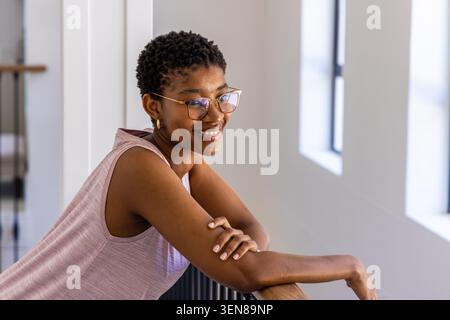 Afrikanische Frau, die sich auf das Geländer lehnt, lächelt im Flur mit Fenstern, trägt goldene Gläser, rosafarbene Oberseite Stockfoto