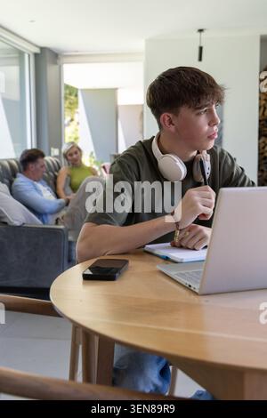 Familie mit Sohn im Teenageralter, der an einem runden Holztisch sitzt und auf einem Laptop mit Kopfhörern studiert Stockfoto