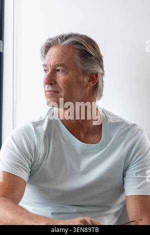 Reifer Mann, der in hellem Raum neben einem hohen Fenster sitzt, ein blassblaues T-Shirt trägt und eine Brille berührt Stockfoto