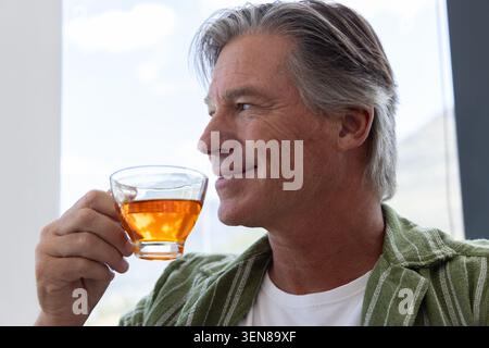 Ein Mann mittleren Alters hält eine Teetasse aus Glas und blickt links aus dem Fenster mit einem grün gestreiften Overshirt Stockfoto