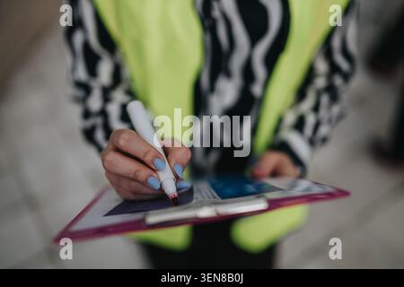Die Person füllt das Formular mit einem Marker auf dem Clipboard aus, während sie eine Warnweste und ein gestreiftes Hemd trägt Stockfoto