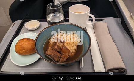 Luxuriöses Business-Class-Essen an Bord mit asiatischer Rindfleischnudelsuppe, frischem Brötchen und heißem Getränk auf einem Tablett-Tisch im Flugzeug Stockfoto