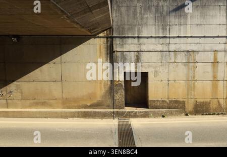 Schmalbeton-Blockwand einer durch Schatten geteilten Straßenunterführung. Bürgersteig und Asphaltstraße davor. Hintergrund für Kopierbereich Stockfoto