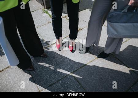 Geschäftsfrauen stehen draußen in Heels auf einem sonnigen Bürgersteig, eine trägt rote Schuhe Stockfoto