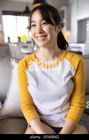 Asiatische Frau, die sich auf der beigen Couch im hellen Wohnzimmer mit einer Vase mit gelben Blumen entspannt Stockfoto