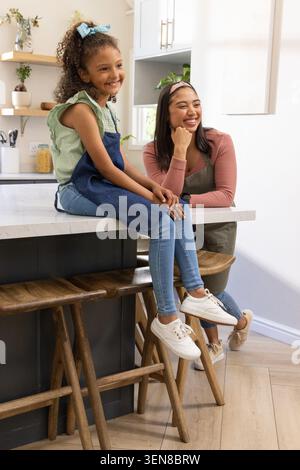 Verschiedene weibliche Mutter und Kind lächeln, sitzen auf Marmor-Arbeitsplatte in der Küche mit Pflanzen Stockfoto