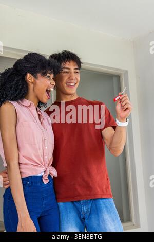 Ein vielseitiges Paar feiert den Empfang neuer Schlüssel und hält den roten Schlüssel im Eingangsbereich Stockfoto