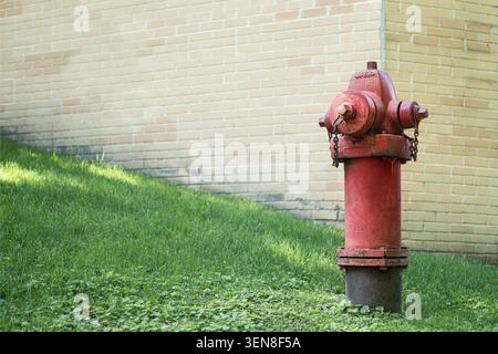 Verwitterter roter Hydrant auf grünem Gras in der Nähe einer Ziegelwand mit Kopierraum auf der linken Seite. Städtisches Infrastrukturkonzept für den Brandschutz, entstehen Stockfoto