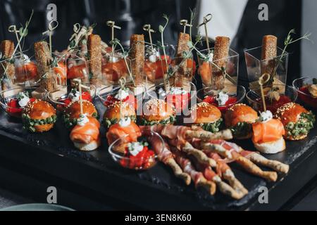 Gourmet-Vorspeisen glänzen auf einem schwarzen Tablett. Burger, Käse und geräucherte Fleischgerichte stehen in kleinen Schüsseln. Holzspieße halten Happen und sind bereit zum Genießen. Fi Stockfoto