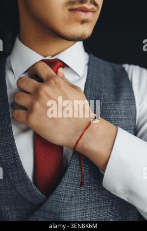 Der Mann passt die rote Krawatte mit konzentrierter Ruhe an. Die Hand liegt sanft auf dem Stoff auf und zeigt dabei Sorgfalt in Gesten. Das Armband aus rotem Faden verleiht dem Benutzer einen dezenten, persönlichen Touch. Profe Stockfoto