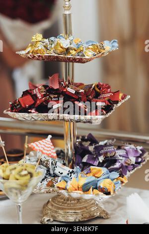 Goldene Süßigkeiten schmücken den silbernen Ständer. Bunte Süßigkeiten leuchten unter warmer Tischleuchte. Die Glasschale bietet Platz für Obst und cremiges Dessert. Elegantes Display Inv Stockfoto