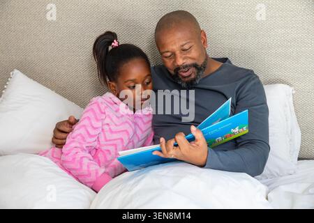 Afroamerikanischer Mann mit dunklem Hemd, das Buch hält, ein Mädchen mit rosa Reißverschluss, das sich auf das Bett stützt Stockfoto