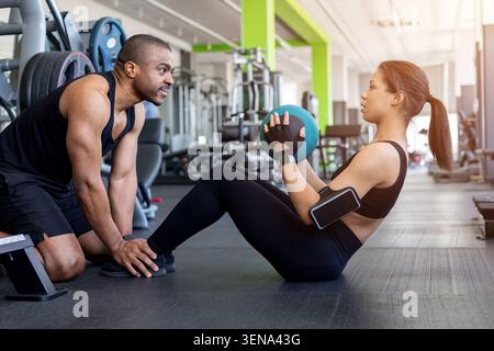 Männlicher Trainer, der Frau beim Sit-up-Training mit Medizinball im Fitnessstudio hilft Stockfoto