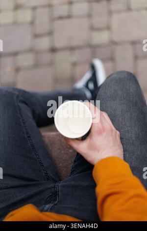 Mann, der auf städtischen Stufen sitzt und eine leere Kaffeetasse aus Papier in lässiger Streetwear hält, mit Jeans, orangefarbenem Sweatshirt und Sneakers, die eine ruhige Pause vermitteln Stockfoto