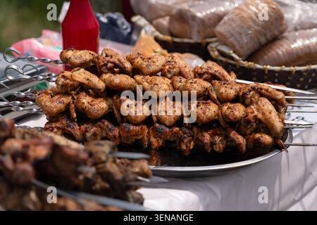 Nahaufnahme gegrillter Hähnchenflügel auf Metallspießen auf einer runden Metallplatte an einem Verkaufsstand im Freien, mit Brotkörben und einer Flasche Stockfoto