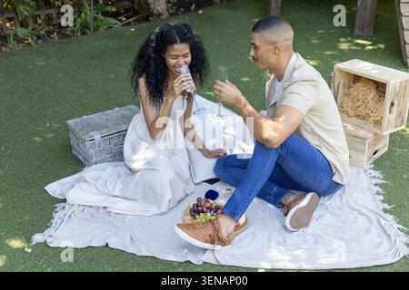 Paar in weißem Kleid, beigefarbenes Hemd, das Flöten auf grauer Decke klirrt, Ringkasten im Garten sichtbar. Romantik, Picknick, Garten, Sonnenlicht, Lachen, Toasts, g Stockfoto