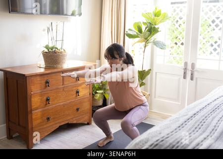 Asiatische Frau, die Squat auf Yogamatte im Schlafzimmer bei Zimmerpflanze in rosa Oberteil, lila Leggings Stockfoto